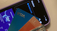Smartphone mit geöffneter Wallet-App, darin eine Bankkarte und ein digitales Veranstaltungsticket. Vor dem Smartphone liegen vier Plastikkarten: eine Visa-Karte, eine Büchereikarte und eine Gesundheitskarte.