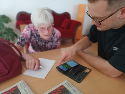 Auf dem Bild sind ein junger Mann und eine Seniorin zu sehen, die zusammen an einem Tisch sitzen und gemeinsam auf ein Smartphone schauen. 