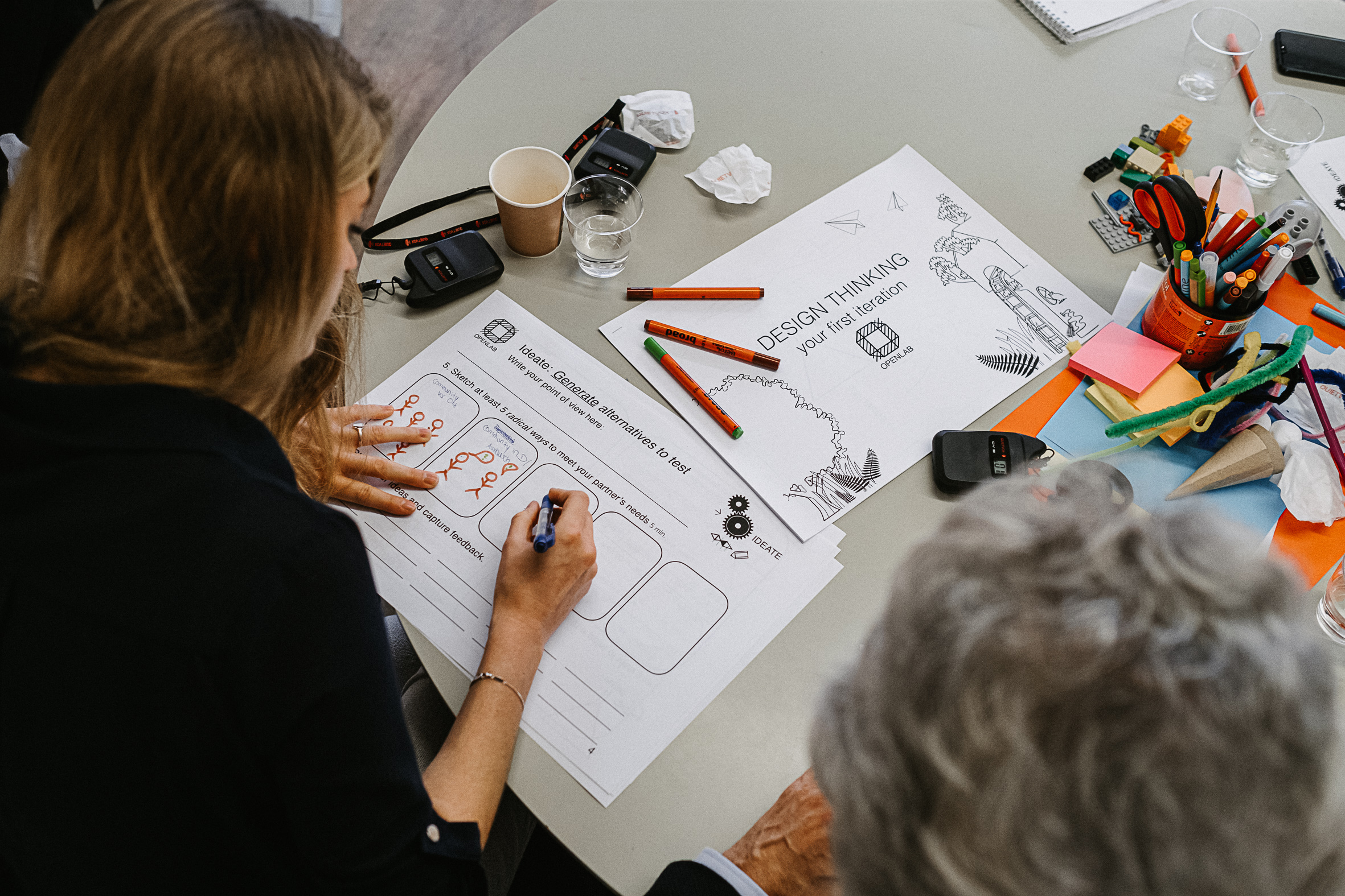 Eine Frau schreibt ihre Gedanken auf ein Blatt Papier. Auf dem Tisch liegt eine Vorlage für ein Design-Thinking-Arbeitsblatt, daneben liegen bunte Stifte verteilt. Eine weitere Frau schaut ihr über die Schulter.